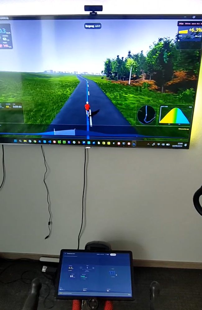 A trainer in front of a wall-mounted TV. The TV shows a game rendering
                a road through trees with a rider on it and a gradient of +6.3 % in the
                corner. On a tablet mounted to the handlebars below, the Indoor Bike app
                shows the same 6.3 % alongside live power, cadence and heart rate.